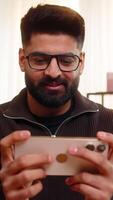 Indian man at home looks at smartphone and celebrates with a big smile and clenched fist after receiving good news. Arabian guy sits alone on sofa, expressing joy and excitement with cheerful gesture video