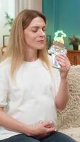 Pregnant middle-aged woman at home eats dark chocolate smiles softly rubs belly enjoying sweet treat calm. Caucasian girl on sofa feels happy boosts mood dark cocoa offers tiny joy while waiting baby. video