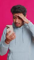 Upset sad Indian man use smartphone typing browsing, play game, loses, surprised by sudden lottery results bad fortune loss fail unlucky news. Arabian Hindu bearded guy isolated on pink background video