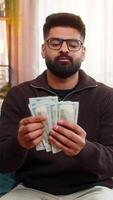 Indian man at home happily counts cash dollars while sitting on sofa, enjoying recent income or lottery win. Hispanic guy expresses joy and financial confidence, celebrating success with satisfaction. video