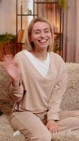 Young adult woman waves hand with a bright smile while sitting on home sofa, greeting with cheerful emotion. Blonde girl expresses friendliness and joy, sharing warmth through open, positive gesture. video