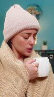 Young woman battling bad cold at home sits wrapped in blanket wearing wool hat and drinks hot tea for relief. Caucasian girl feels weak on sofa breathing and trusting hot tea to restore health soon video