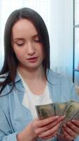 Young woman at home happily sitting on sofa counting large pile stack of dollar cash bills, celebrating wealth. Caucasian girl joyful managing successful personal budget, satisfied financial profit video