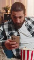 Young man sits at home with popcorn bowl, casually looking at smartphone screen feed in contented ease, watching movie film. Amazed Caucasian guy on sofa shouts wow, discovering something astonishing video