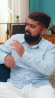 Indian man at home tries dance moves looks unsure then laughs embarrassed enjoying rhythm. Arabian guy on sofa celebrates fun moment chuckling over practice showing lighthearted spirit and enthusiasm video