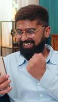 Indian man at home gets amazing smartphone message eyes widen shouts wow celebrates victory pure joy. Arabian guy on sofa raises phone smiles triumph arms up happy cheer winning great online success. video