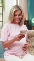 Pregnant middle-aged woman at home smiles typing on smartphone celebrating good news message and lifting victory win hand sign. Happy Caucasian girl on sofa feels anticipation texting on mobile phone. video
