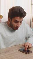Young Indian man with bitcoin checks cryptocurrency exchange charts on smartphone app calculates approximate profits, looking on BTC coins in hand celebrating win. Hispanic guy looks satisfied at home video
