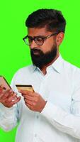 Indian man holds smartphone and credit card while entering numbers, showing online shopping, payment or transaction. Arabian guy isolated on chroma key background expressing digital purchase action. video