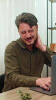 Middle-aged man at home rubs aching neck shuts laptop annoyed by posture pain after marathon work. Young adult guy at table grimaces rubbing muscles tension hopes pain eases after overtime shift soon video