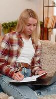 Young adult woman writes in notebook while focusing on laptop screen on home sofa, calculating expenses. Blonde girl manages financial records with care, staying organized and concentrated on task. video