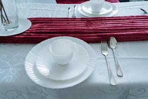 A beautifully arranged dining table showcasing elegant white plates and polished silverware, all complemented by a striking red table runner that adds a vibrant touch to the setting photo