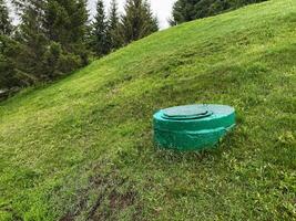 A Green Utility Access Cover is strategically placed on a steeply sloped Green Grass Field that is elegantly framed by a lush Forest, resulting in a truly picturesque and idyllic countryside setting photo