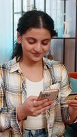 indiano jovem mulher às casa sentado em sofá com Smartphone entrando crédito cartão detalhes, alegremente compras on-line. árabe feliz menina satisfeito, sorridente durante bem sucedido comprar, desfrutando comércio eletrônico video