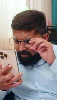 Indian man at home wearing glasses squints reading smartphone text holding screen closer focused. Arabian guy on sofa leans forward eyes narrow tries seeing message shows effort concentration vision. video