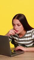 Woman sitting at table attentively reading complex material on laptop with focused expression during online learning. Girl on yellow background shows concentration engagement in serious study process video