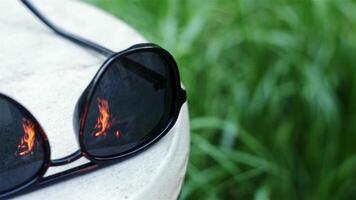 Close-up of sunglasses in which burning big fire and bicycle are reflected against background of green grass in nature during day in daylight. video