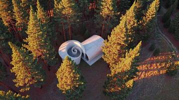 Aerial view capturing a unique spiral shaped concrete house nestled in a dense pine forest, glowing warmly in the sunset, highlighting innovative architecture harmonizing with nature video
