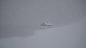 Heavy snow falling and strong wind blowing around an isolated mountain chalet with red roof during a winter blizzard, creating a dramatic and captivating scene of nature's power video