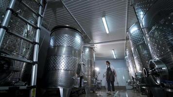 A happy successful farmer or winemaker is walking in the middle of wine tanks and checking a fermentation grade of high quality wine production in a modern winemaking facility or winery. 3 video