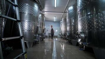 A happy successful farmer or winemaker is walking in the middle of wine tanks and checking a fermentation grade of high quality wine production in a modern winemaking facility or winery. 2 video