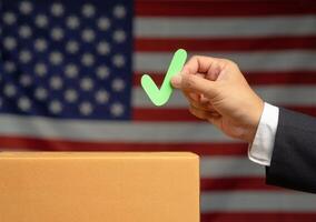 Hand holding a green check mark symbol overhead the voting box at place election with the American flag background photo