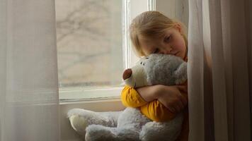 Sad little girl child sitting looking at window hugging teddy bear favorite toy childhood home unhappy kid daughter problem alone crisis pensive thinking mental health psychology stress upset suffer photo