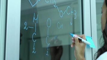 Close-up of female scientist writing chemical formulas on glass board with blue marker in laboratory. video