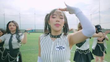 A group of dancers performing on a grassy field, wearing stylish outfits with a sporty vibe. The main dancer is in focus, showcasing a confident pose, while others are in the background video
