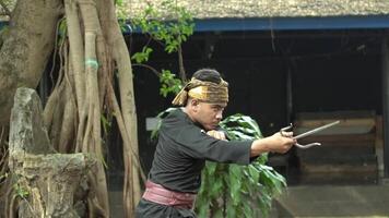 A person performing a traditional dance or martial arts move outdoors, wearing a black outfit and a decorative headband. The background features lush greenery and a rustic setting. video