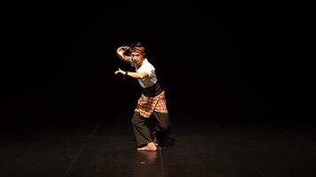 A dancer performing on a dark stage, showcasing traditional movements. The dancer wears a traditional outfit with a checkered sarong and a headband, emphasizing cultural expression. video
