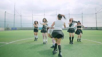 A group of six dancers performing on a green turf field, dressed in stylish black and white outfits. They are in dynamic poses, showcasing their choreography under a cloudy sky. video