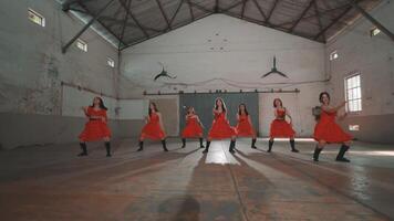 A group of six dancers in red dresses performing in an industrial space. The dancers are synchronized, showcasing dynamic movements with a spotlight illuminating them. The background features a rustic video