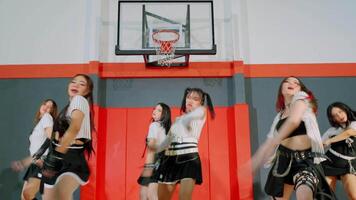 A group of dancers performing in a gymnasium, showcasing energetic choreography. The lead dancer, wearing a striped top and skirt, strikes a dynamic pose. The backdrop features a basketball hoop. video