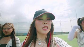 A dynamic scene featuring young women dancing energetically on a sports field. The focus is on one woman in the foreground wearing a cap and layered jewelry, with others in the background video