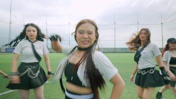 A group of six female dancers performing on a green turf field. They are dressed in stylish outfits, showcasing dynamic dance moves against a cloudy sky backdrop. video