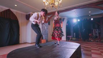 A couple performing a traditional dance on stage in a festive setting. The woman wears a colorful dress with floral patterns, while the man is in a white shirt and black pants video