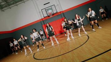 A group of young dancers performing in a gymnasium. They are dressed in coordinated outfits, showcasing a choreographed routine. The basketball hoop in the background adds to the sports theme. video