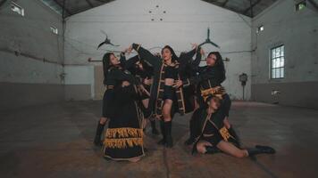 A group of dancers performing synchronized movements in an industrial space. The dancers are dressed in black outfits with yellow accents, showcasing dynamic poses and energy video