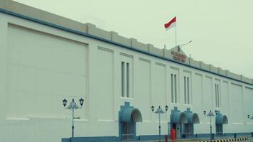 A large, white building with a flat roof and decorative elements. The Indonesian flag is prominently displayed on top. Street lamps line the sidewalk in front of the building video