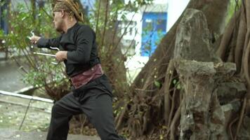 A martial artist performing a dynamic move in a natural setting with large trees and roots in the background. The individual is dressed in black attire and a red belt, showcasing agility and focus. video