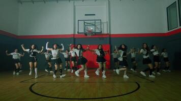 A group of dancers performing a choreographed routine in a basketball gym. They wear matching outfits and showcase synchronized movements, with a basketball hoop in the background. video