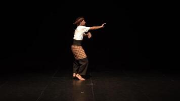 A dancer performing on stage with a black background, showcasing a dynamic pose. The dancer wears traditional attire, emphasizing movement and expression. video