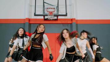 A dynamic dance performance by a group of five female dancers in a gymnasium. They wear stylish outfits with chains and perform synchronized moves in front of a basketball hoop. video