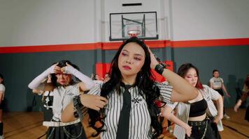 A group of dancers performing in a gymnasium, featuring a central female dancer with long black hair and striking makeup. The dancers wear matching striped outfits and showcase energetic choreography. video