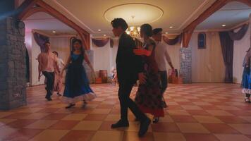 A lively dance scene in a spacious hall with a checkered floor. Several couples are dancing, showcasing traditional attire. Soft lighting enhances the festive atmosphere. video