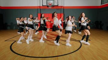 A group of dancers performing a choreographed routine in a basketball gym. They are wearing stylish outfits and showcasing energetic movements, with some dancers in the foreground video