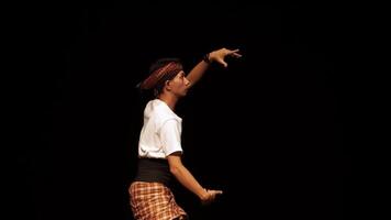 A young dancer performing on stage, showcasing traditional dance moves. The dancer wears a white shirt, patterned pants, and a headband, with a dramatic spotlight highlighting their movements a video