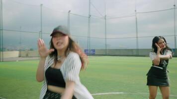 Three young women performing a dance routine on a sports field under a cloudy sky. They wear stylish outfits, including caps and sporty attire, showcasing energy and enthusiasm. video