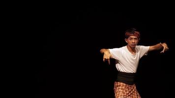 A young male dancer performing on stage, wearing a traditional outfit with a headband. The background is dark, emphasizing the dancer's movements and expressions. video